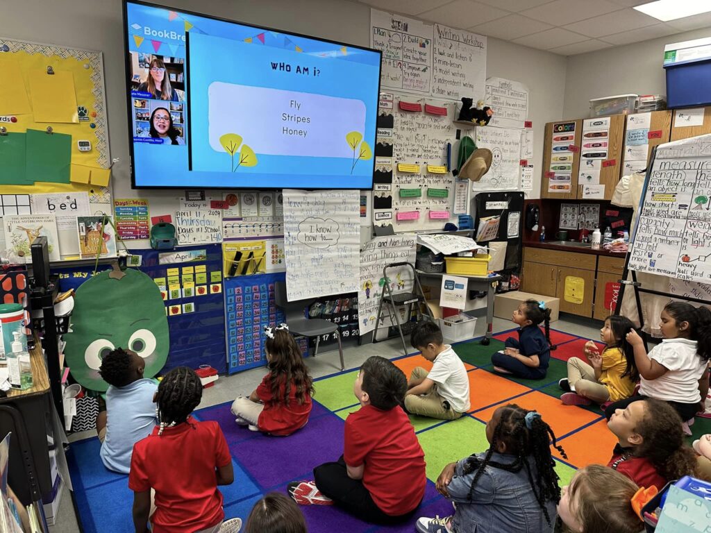 A photo of K-1 Graders sitting on a colorful carpet of squares in a classroom watching a virtual author talk with author Kate Messner